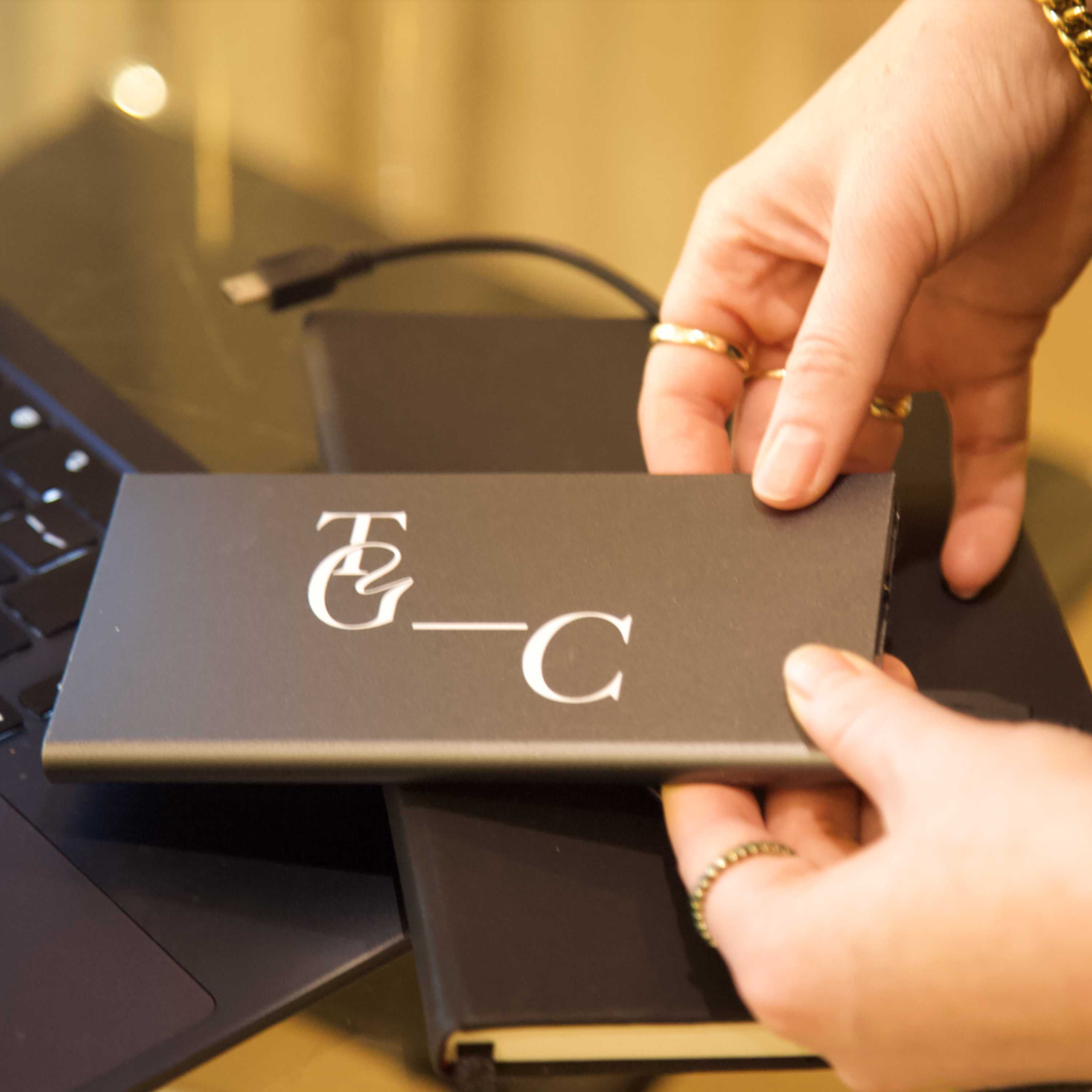 Person holding a power bank with TGC initials against a blurred background with a notebook. 