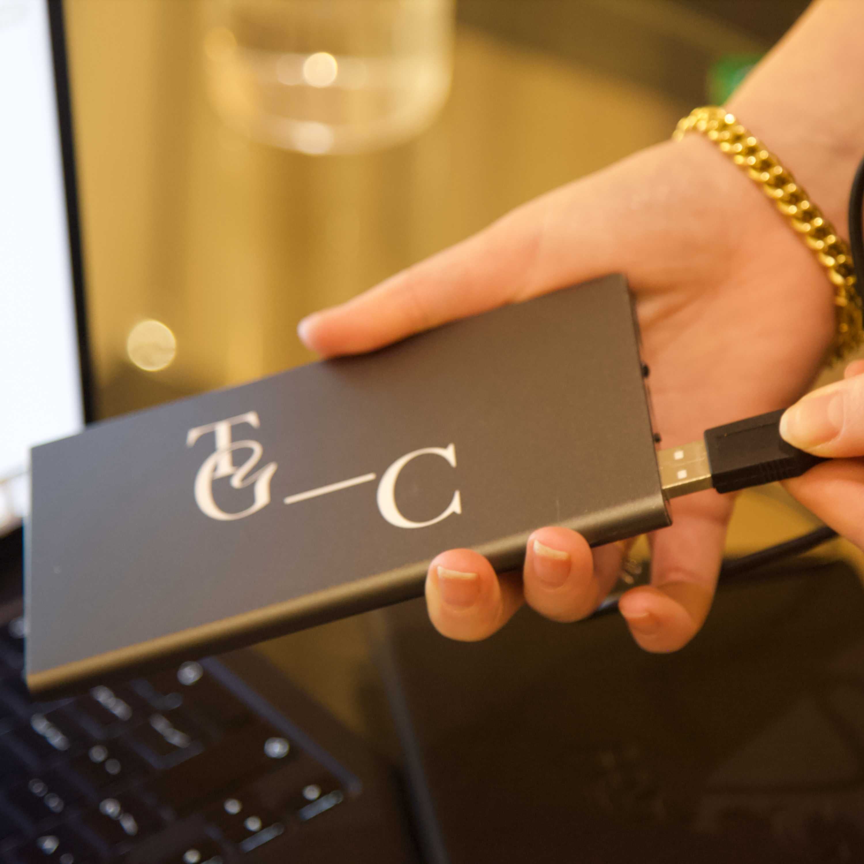 Hand holding a Power Bank with the TGC logo on in front of a laptop.