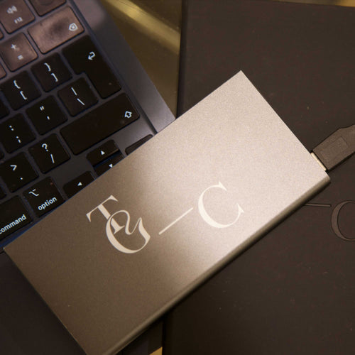 Metallic power bank with TGC logo on a dark surface next to a laptop.